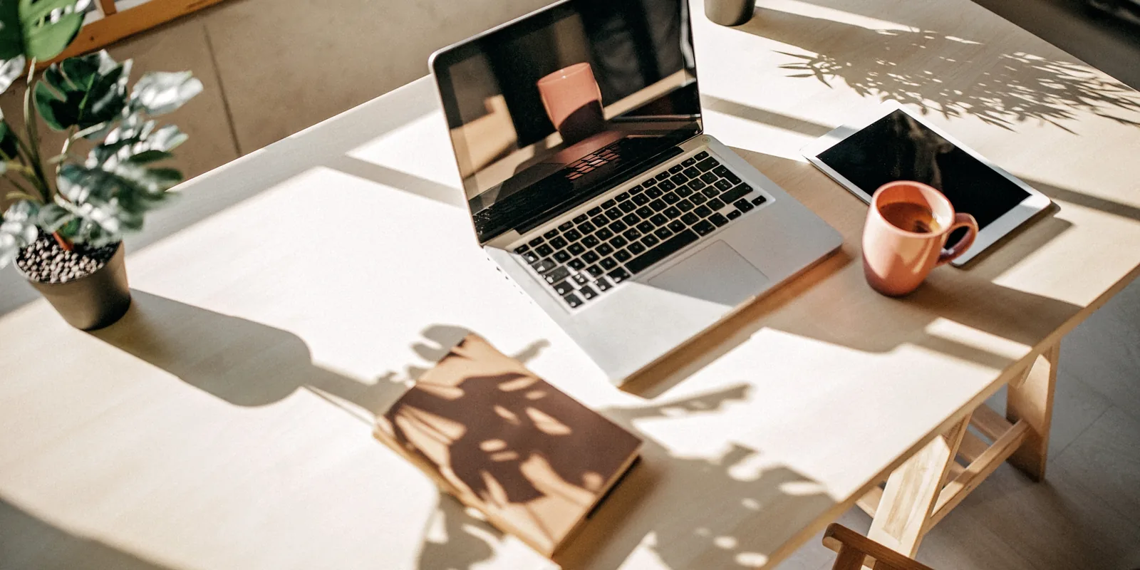 Sonniger Schreibtisch mit aufgeklapptem Laptop, Tablet, Kaffeebecher und Notizbuch, Alltagssituation vor der Wahl des passenden KI-Tools